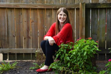 Genç kadın bahçesinde dinleniyor. Arka planda ahşap çitler var ve çiçekler açıyor. Siyah pantolon ve kırmızı bluz giyiyor..