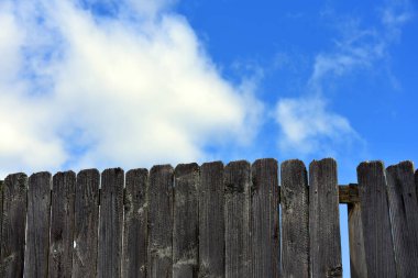 Arkaplan görüntüsü, paslı, pürüzlü ahşap bir çitin yakın çekimini gösteriyor. Mavi gökyüzü ve bulutlar arka planı oluşturur.