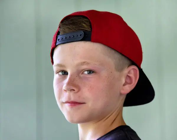 stock image Young boy looks at camera with his baseball cap turned backwards.  He has a small smile on his face.  Freckles sprinkle across nose and cheeks.  Cap is red and black.