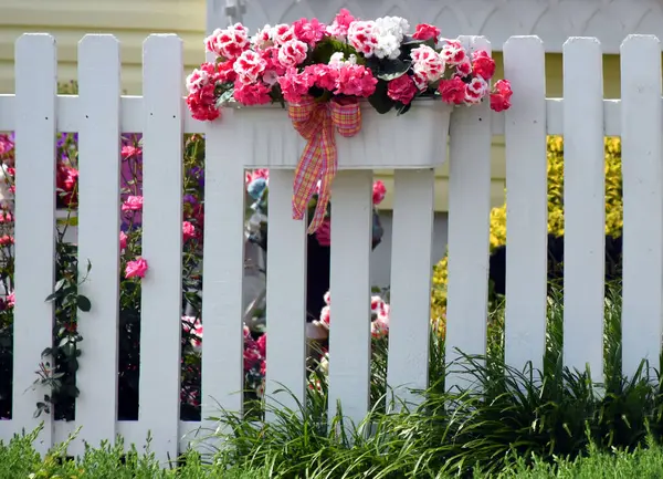 Pink silk flowers fill white wooden box.  It hangs on white wooden fence surrounding yard.  Real pink roses peek between slats.
