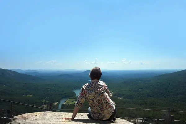 stock image Visitor sits absorbing the view at the top of Chimney Rock, Chimney Rock State Park, North Carolina.  Visitor is a senior citizen and is wearing jeans and print shirt.