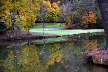 Autumn trees are reflected in the still waters of Steele Creek Lake in Bristol, Tennessee.  Golf cart track curves around golf course. clipart