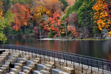 Bays Mountain Lake in Bays Mountain Park, Kingsport, Tennessee is showing its' colors.  Autumn reflects in calm surface of lake, and concrete dam and path runs in front of lake. clipart