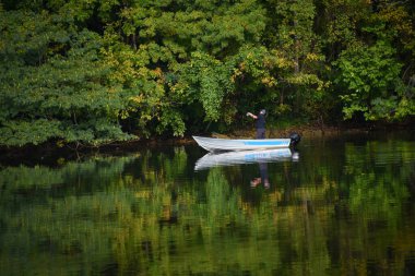 Man casts his rod in the hopes of catching a fish.  He is standing, alone, in his boat on a the shores of Duck Island, Warriors Path State Park. His reflection shows in the still waters and green foliage. clipart