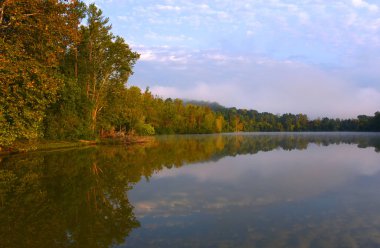 Trees and sky are reflected in the calm water of Watts Lake, Soaring Eagle Campground.  Sunrise brightens sky and tints trees with red and gold. clipart