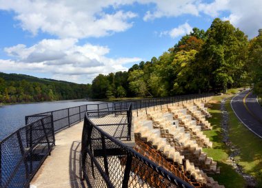 Winding road leads to Bay Mountain State Park.  It passes besides the Dam and waterfall.  Concrete dam is topped with viewing area protected by black metal rail. clipart