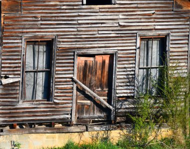 Front shows old home that is leaning slightly.  Exterior has boards that are weathered and faded.  Doors are uneven and faded with age. clipart