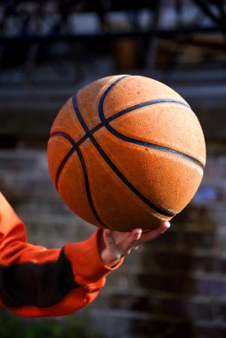 Basketball shows worn places where it has been dribbled on concrete outdoors.  Boy holds basketball in one hand. clipart