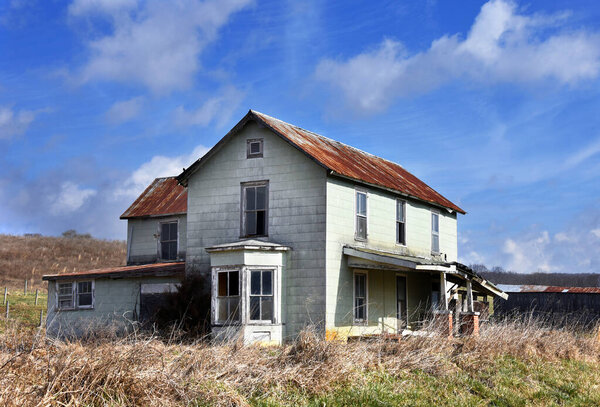 Очаровательный загородный дом заброшен. Крыша крыльца рушится, окна разбиты. Двухэтажное жилище имеет ржавую железную крышу.