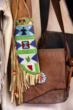 Long, beaded Indian knife sheath is trimmed with leather fringe and hngs on a wall with leather bags and pipe. clipart