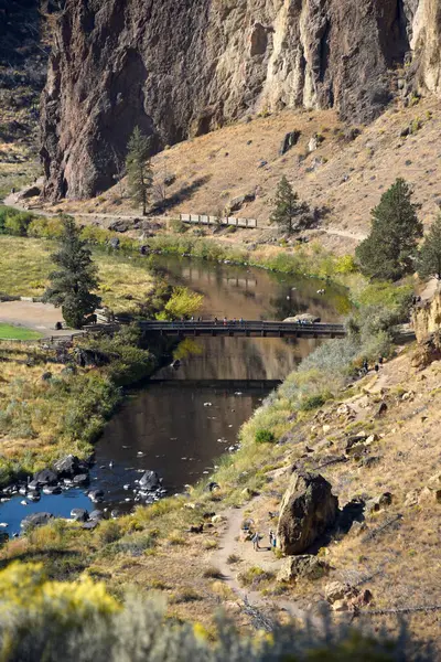 Ziyaretçiler Smith Rock State Park, Oregon 'da yaya köprüsünden geçiyorlar. Yüksek açı, Eğri Nehir 'i ve dağ tabanının etrafındaki izleri gösteriyor..