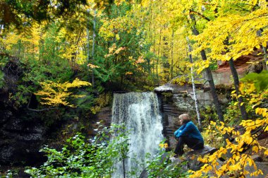Yetişkin kadın Michigan 'daki Keweenaw Yarımadası' nda Macar Şelalesi 'nin önünde dinleniyor. Sonbahar sarısı, yürüyüşçüyü düşünceleri ve şelaleyle çevreledi..