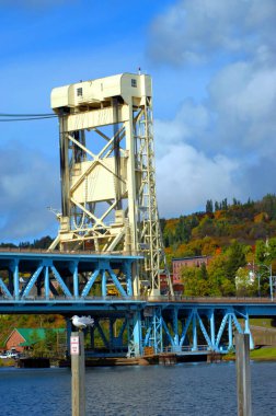 Houghton ve Hancock, Michigan 'ı birbirine bağlayan Portage Lake Lift Köprüsü' nün uzun kulesi beyaza boyanmıştır ve asma köprü maviye boyanmıştır. Martı iskelede oturuyor ve sonbahar renkleri manzarası.