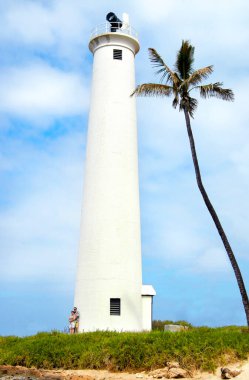 Couple stands at the base of Barber's Point Lighthouse to show comparison of size.  Lone palm tree leans toward lighthouse, which stands on Oahu in Hawaii.                              clipart