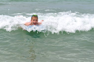 Küçük çocuk Florida 'daki Madeira Sahili' nde küçük bir dalgada sörf yaparken gülüyor. Kafasında yeşil şnorkel gözlüğü var..