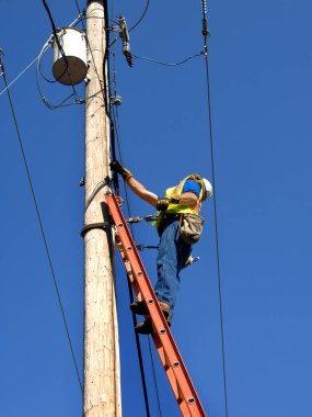 Çizgi hakemi sorunu çözmek için elektrik direğine tırmanır. O bir kask ve emniyet kemeri takarak bir merdivenin üzerinde duruyor. Mavi gökyüzü onu çerçeveler..