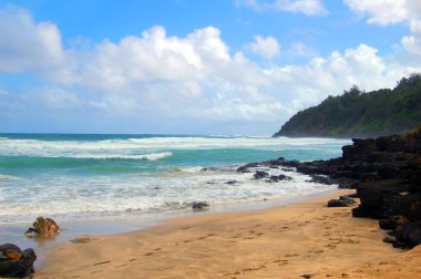 Hawaii, Kauai Adası 'ndaki terk edilmiş kayalık ve kumlu sahil, yumuşak turkuaz dalgalarla yıkanır. Adanın kıvrımları uzaklardan görülüyor.