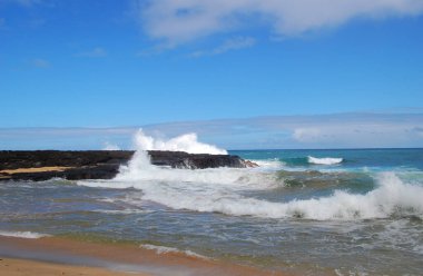 Güzel mavi gökyüzü ve turkuaz su, Hawaii 'nin Kauai adasındaki bu siyah lav kayasının etrafını sarar. Dalgalar kayalara çarpar.