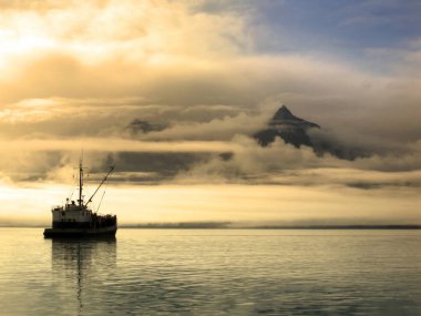 Ship sits quietly on the Bay of Valdez, while sunrise colors the clouds and mist over the Chugach Mountains in Alaska.  Sky begins to clears in right corner of image. clipart