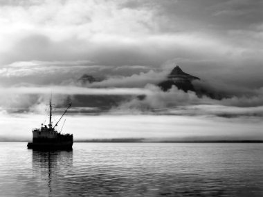 Ship sits quietly on the water while sunrise colors the clouds and mist over the Bay of Valdez in Alaska.  Sky clears in right corner of image. clipart