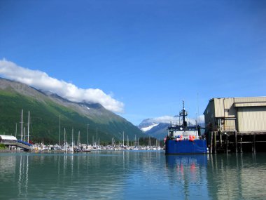 Mavi balıkçı teknesi Valdez, Alaska limanında bir rıhtıma yanaştı. Körfezin karşısındaki küçük tekne limanı görülebilir. Chugach Dağları arkaplanda yükseliyor.