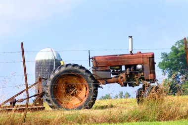 A relic of farming is parked at the fence row to be sold.  Tractor and bushhog stands idle and rusting.  A lone silo stands in the background. clipart