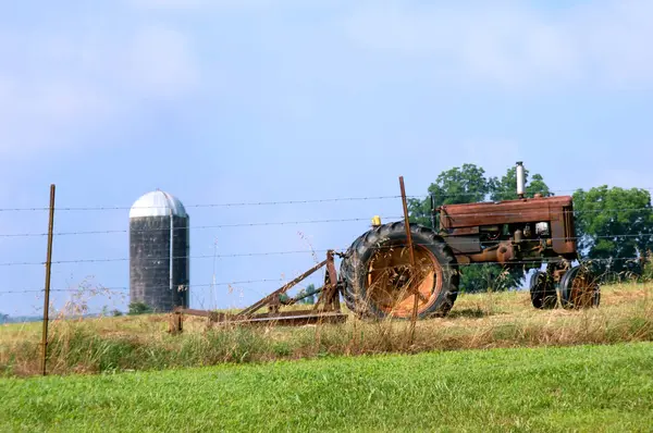 stock image Vintage tractor and mower sit idle besides a barbed wire fence.  Silo and blue sky fill background.