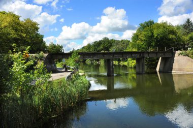 Man pulling a wagon and child disappears around the bend in a road under railroad track.  Track travels over Yahara River in Stoughton, Wisconsin. clipart