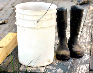 Ahşap verandadaki 5 galon kovanın yanında çamurlu iş botları. Veranda tahtaları çatlamış ve soyulmuş. İş eldivenleri arka plandaydı. Çizmeler bir iş etiğini temsil ediyor.