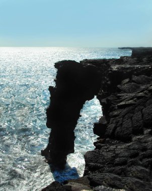 Hawaii Volkanları Ulusal Parkı 'ndaki Kraterler Zinciri' nin sonundaki Holei Deniz Kemeri akşam güneşiyle siluete bürünmüştür. Güneş ışığı ufukta parlarken su kemerin tabanında kıvrılır..