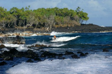 Genç adam sörf tahtasında dengede duruyor ve Hawaii 'nin büyük adasındaki Ahalanui Parkı yakınlarındaki engebeli kayalardan uzak durmaya çalışıyor..