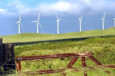 Farm pastureland sustains windpowered turbans.  Rustic metal gate and fence posts aid in fencing off area. clipart