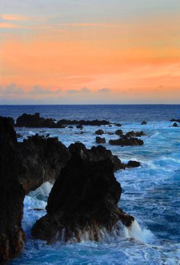 Hawaii 'nin büyük adasında yükselen renkli günbatımı, gökyüzünü turuncuya boyar. Mavi su ufukta buluşur ve dalgalardan fışkırır Laupahoehoe Plaj Parkı 'ndaki siyah lav yığınlarını hafifletir..