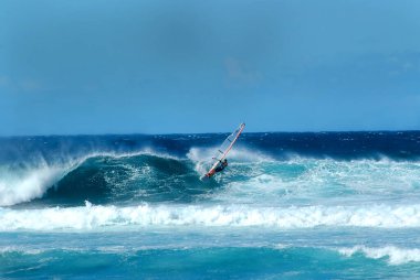 Rüzgâr sörfçüsü Hawaii, Kauai açıklarında büyük bir dalgaya biner. Tahtasının gölgesi ve yelkenleri parlak mavi suları yansıtıyor..                        