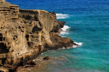 Cliff side of the Green Sand Beach, Big Island of Hawaii, wind and wave erosion is seen in the sand stone cliff face.  Aqua blue water and waves surround cliff. clipart