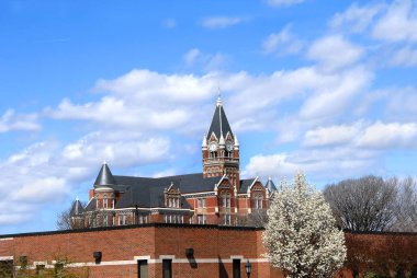 Wichita, Kansas 'taki Friends Üniversitesi' ndeki saat kulesine mavi gökyüzü ve bulutlar çerçevelenmiş. Beyaz bahar çiçekleri süs armudunda patlar.