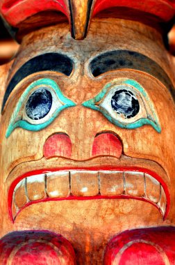 Oyulmuş tiki totem suratını buruşturuyor. Dişler görünüyor ve turkuaz mavi çerçeveli gözler bakıyor..