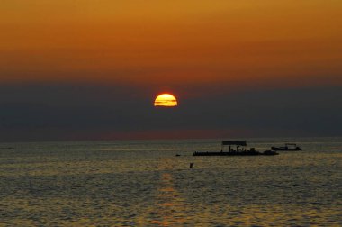 Big Island sunset silhouettes flat bottom boat and skiff.  Sun is a ball of fire and tints sky a bright orange. clipart