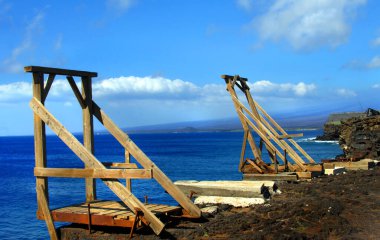 Two wooden canoe hoists perch on steep cliffs edge at South Point on the Big Island of Hawaii.  Vivid blue water  clipart