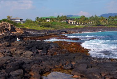 Female tourist sits on the black lava rock near Shipwreck beach.  She is alone and enjoying the view of resort, ocean and mountains. clipart