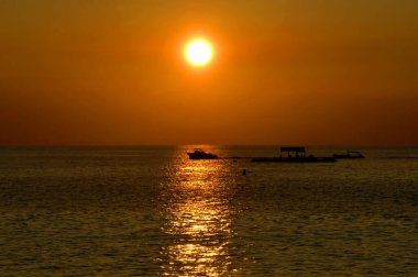 Boater hurries home for his supper.  He crosses the line of reflection from a setting set.  Sun is a ball of orange fire against an orange tinted sky on Big Island. clipart