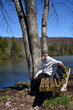 Male teenager sits on a wooden bench besides Bays Mountain Lake in Kingsport, Tennessee.  He has on a hoodie and his hat is backwards. clipart