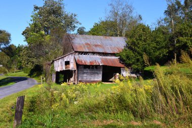 Aging country barn sits at edge of a curving road.  Hay is stored in loft, but weeds overgrown pasture.  Tin roof is rusting and boards are broken and rotting. clipart