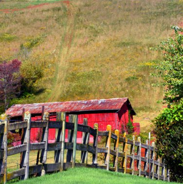 Rustic wooden fence runs in front of a red, wooden barn in Tennessee.  Barn has rusting roof, and sits at the foot of a mountain.  Dirt track leads up mountain. clipart