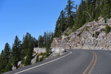 Bisikletçiler Krater Gölü Ulusal Parkı 'ndaki Rim Drive' da yokuş yukarı tırmanıyorlar. Krater Gölü etrafındaki virajlı yolda ilerliyorlar..