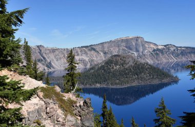 Büyücü Adası, kül konisi Oregon 'daki Krater Gölü' nün cam suyuna yansıyor. Manzara mavi gökyüzünü ve derin mavi suyu gösterir.