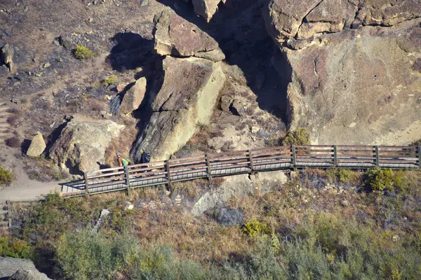 Oregon 'daki Smith Rock Eyalet Parkı' nda iki dağcı yaya köprüsünden geçiyor. Tırmanma noktasına yürürken sırtlarında ekipman taşıyorlar..