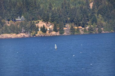 Man rides the wind as he windsurfs on the Columbia River, in Oregon.  He is alone on the blue water.  Train travels the shore line behind. clipart