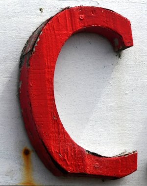 Büyük, kırmızı ahşap C beyaz reklam panosuna asılır. Mektup yıpranmış ve soyulmuş..  