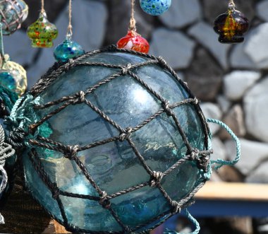 Büyük bir el patlaması, cam şamandıra dışarıda güneşin altında duruyor. Cam su renginde ve düğümlü ipe sarılı..